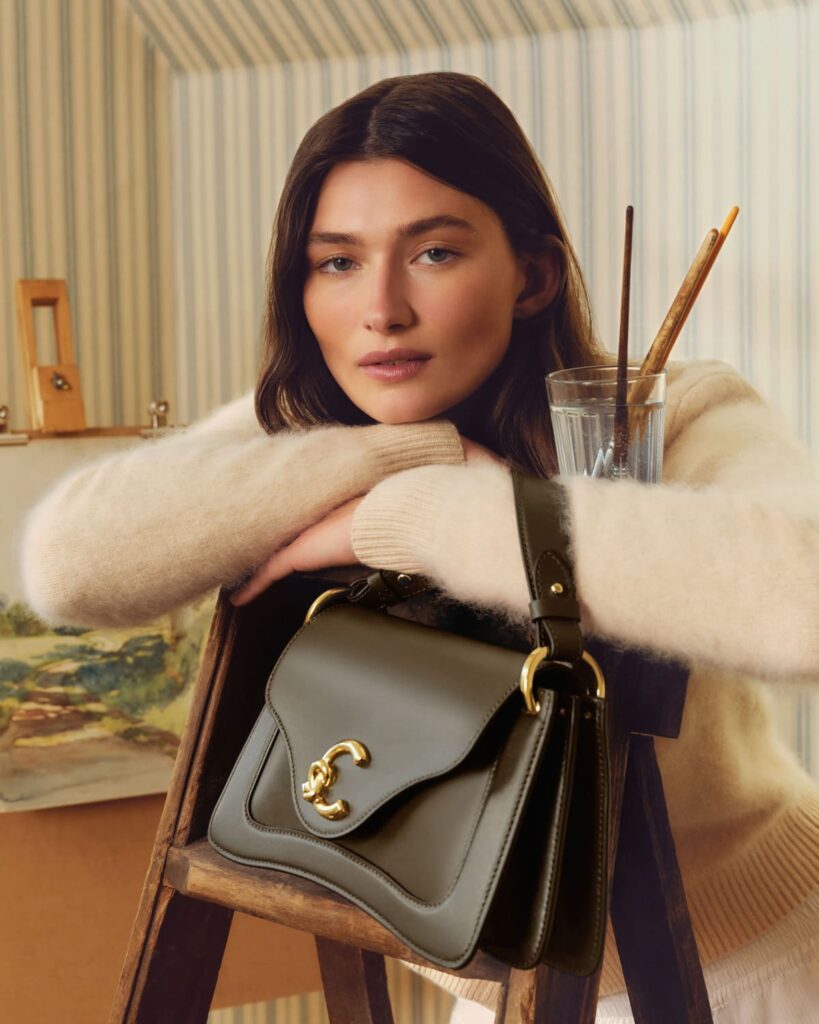 Modella con una borsa Coccinelle presumibilmente C-ME Lock nell'ambientazione della campagna The Gardener's Studio, autunno inverno 2026