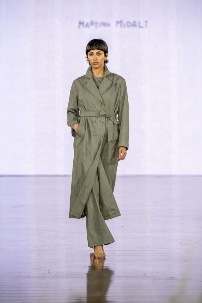 Trench verde Martino Midali con chiusura doppiopetto e cintura in vita, abbinato a pantaloni ampi dello stesso tono.