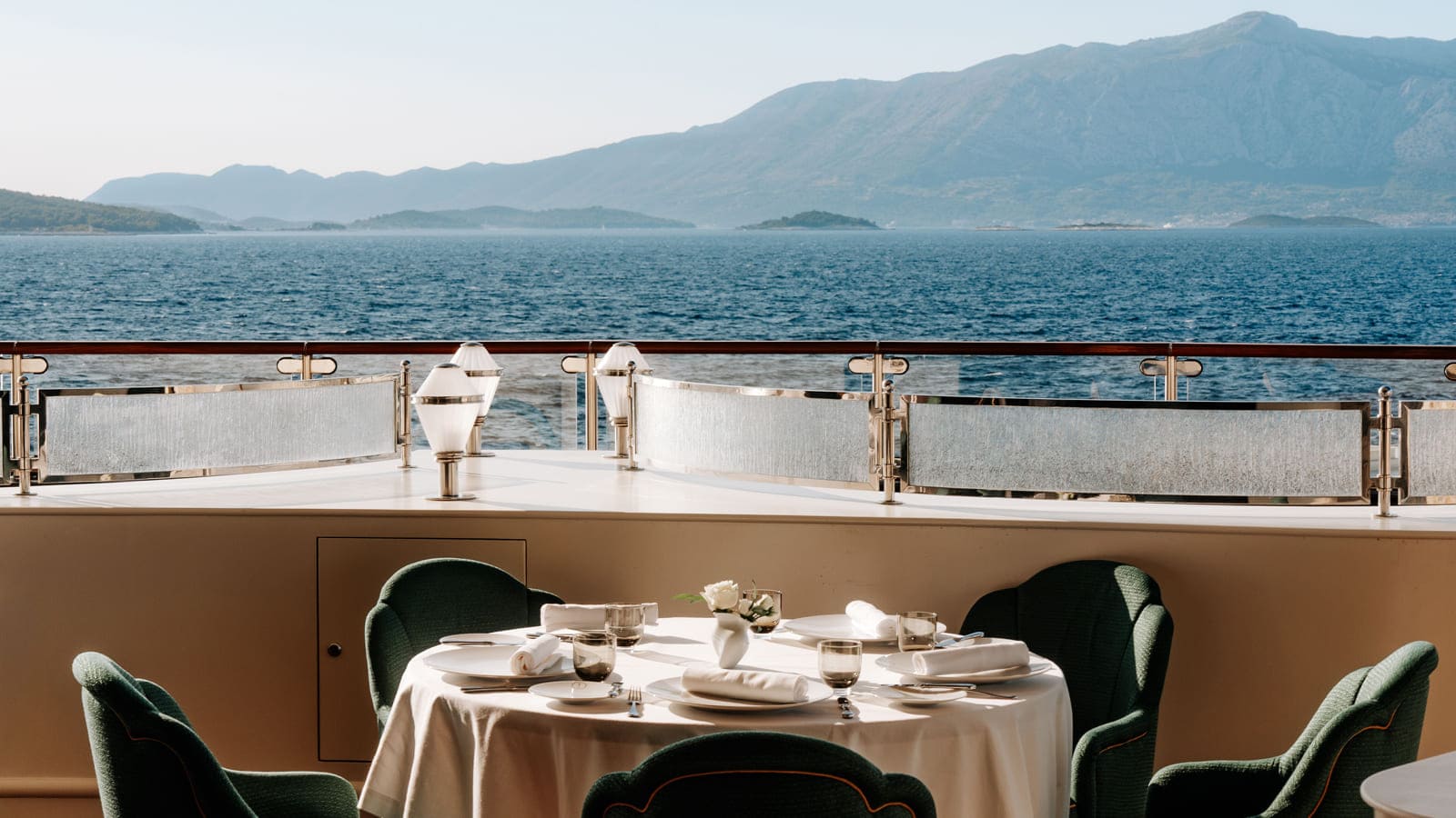 Tavolo apparecchiato e poltroncine verdi in uno spazio ristorazione di Four Seasons I, con vista sul mare e sulla costa.