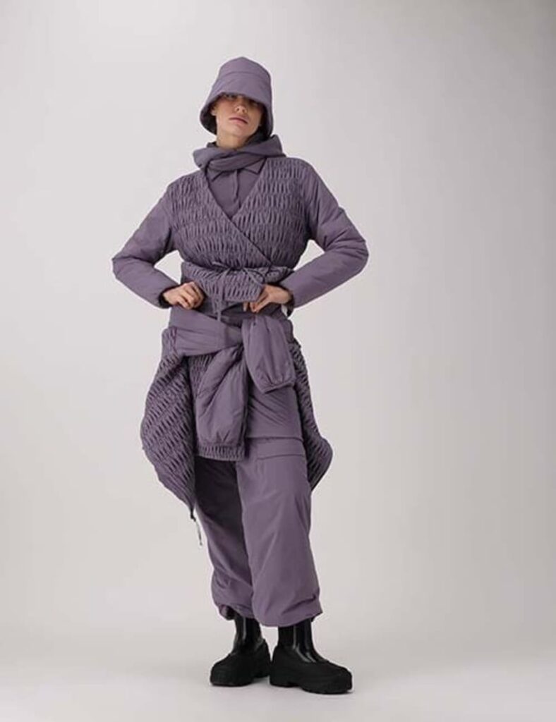 Modella in studio con abbinamento e cappello nei toni del viola, mani sui fianchi e calzature nere.