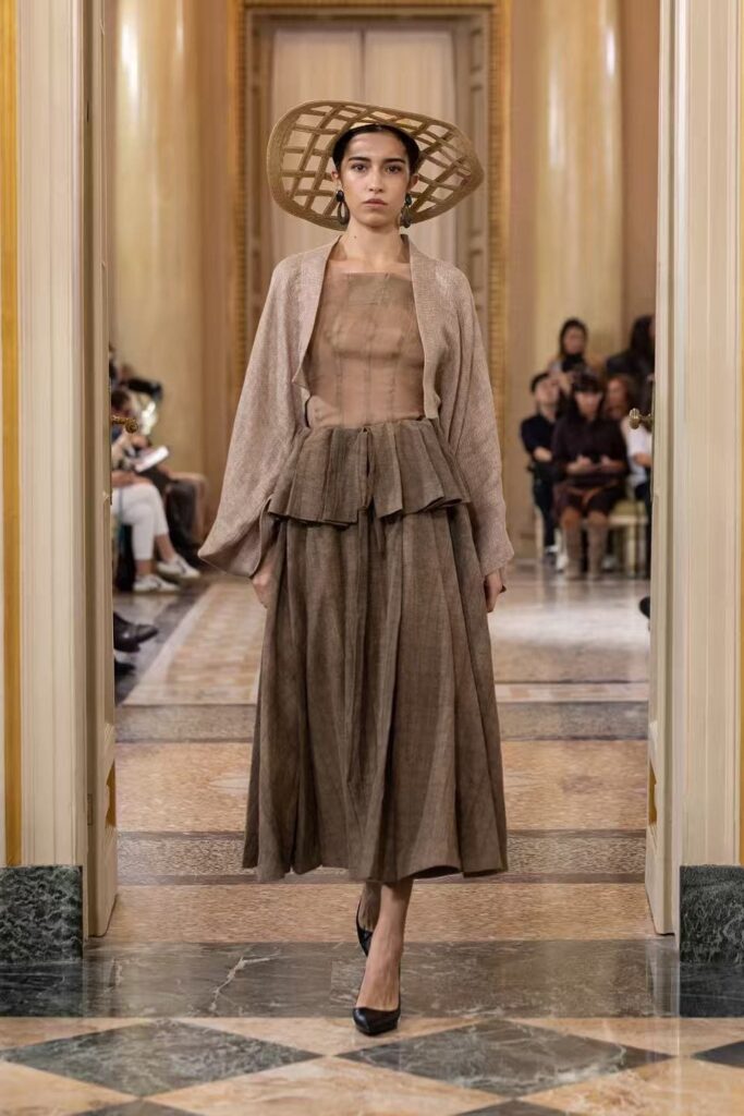 Look Sancailiyi nei toni del marrone, con gonna ampia, giacca aperta e cappello a larga tesa traforata.