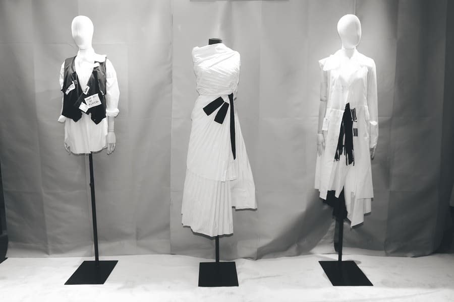 Tre figure espositive con capi Malloni: gilet su camicia, abito drappeggiato con pieghe e sovrapposizioni asimmetriche.