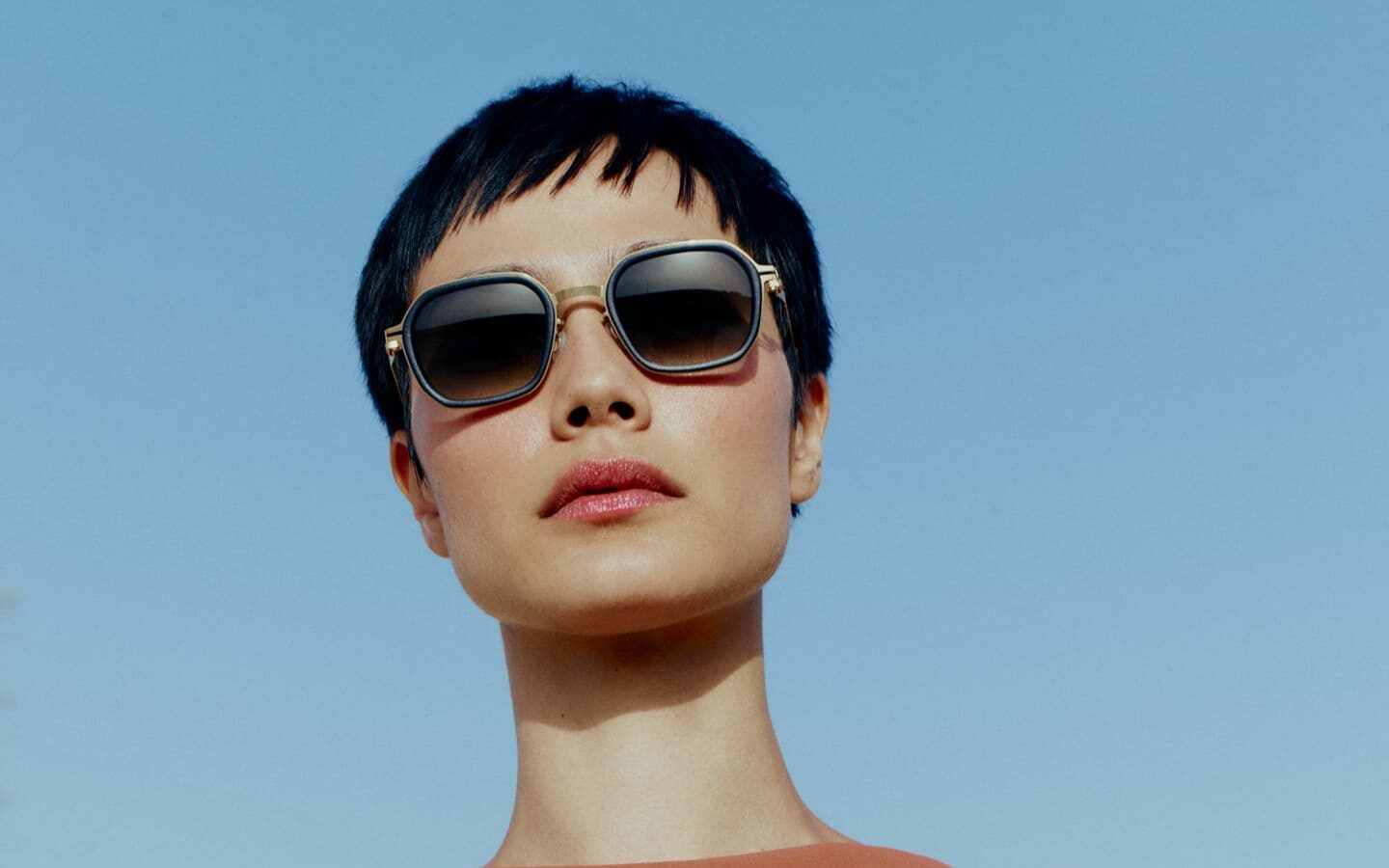 Donna con capelli corti, occhiali da sole geometrici e abito ruggine sotto un cielo azzurro