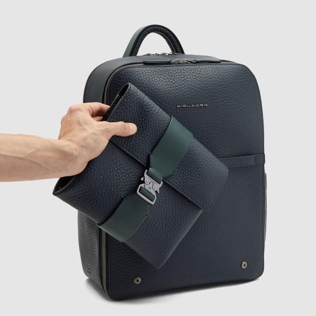 Mano che rimuove la tasca frontale removibile con fascia centrale verde, dallo zaino della collezione Piquadro MATERIA
