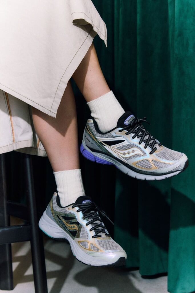Sneakers Saucony Daikanyama in mesh chiaro indossate con calze bianche e gonna chiara, davanti a una tenda verde.