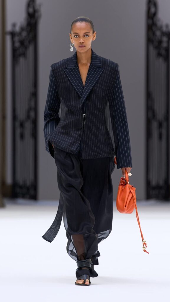Modella con giacca gessata scura, pantaloni trasparenti e piccola borsa arancione