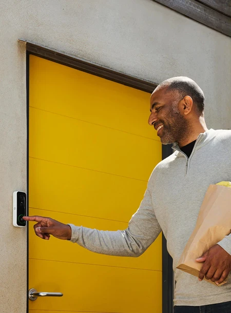 Uomo con un pacco tra le braccia suona il Campanello Smart Yale installato su una porta gialla"