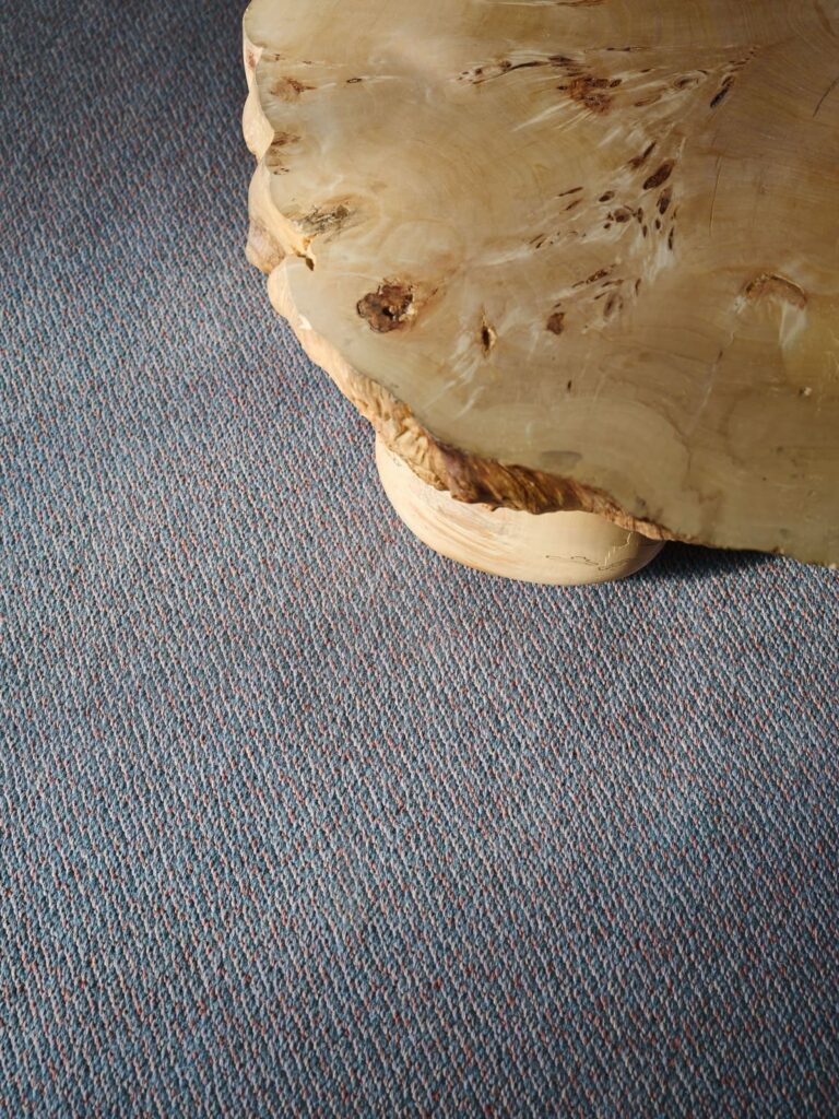 Dettaglio della texture del tappeto Korn della collezione Landskab di Kasthall nella nuova colorazione Malm, con l'angolo di un tavolino in legno