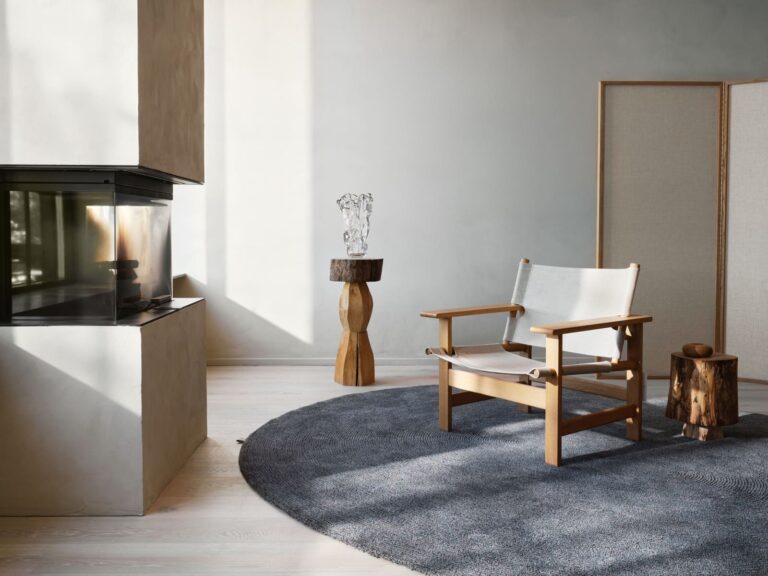 Tappeto ovale Spire Oval della collezione Landskab di Kasthall nella colorazione Malm, in un living con camino e poltrona in legno