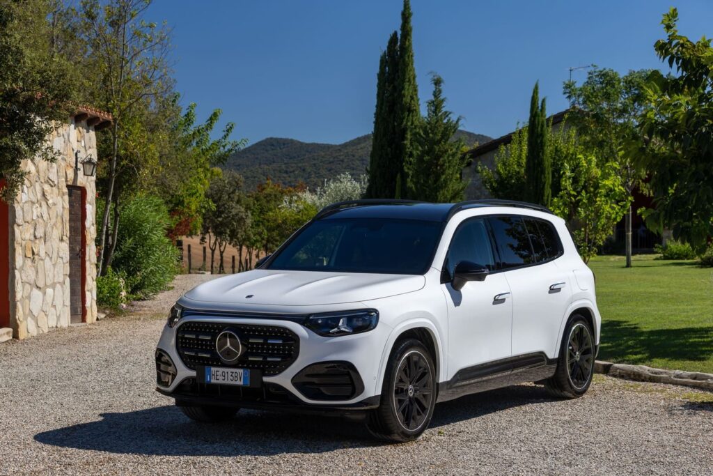 Mercedes GLB bianca vista di tre quarti anteriore su un piazzale in ghiaia, fra alberi ed edifici in pietra.