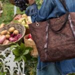 Borsa Campomaggi Bella In The Market indossata a spalla da una donna in denim, tra cesti di frutta e verdura.