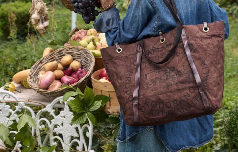 Borsa Campomaggi Bella In The Market indossata a spalla da una donna in denim, tra cesti di frutta e verdura.